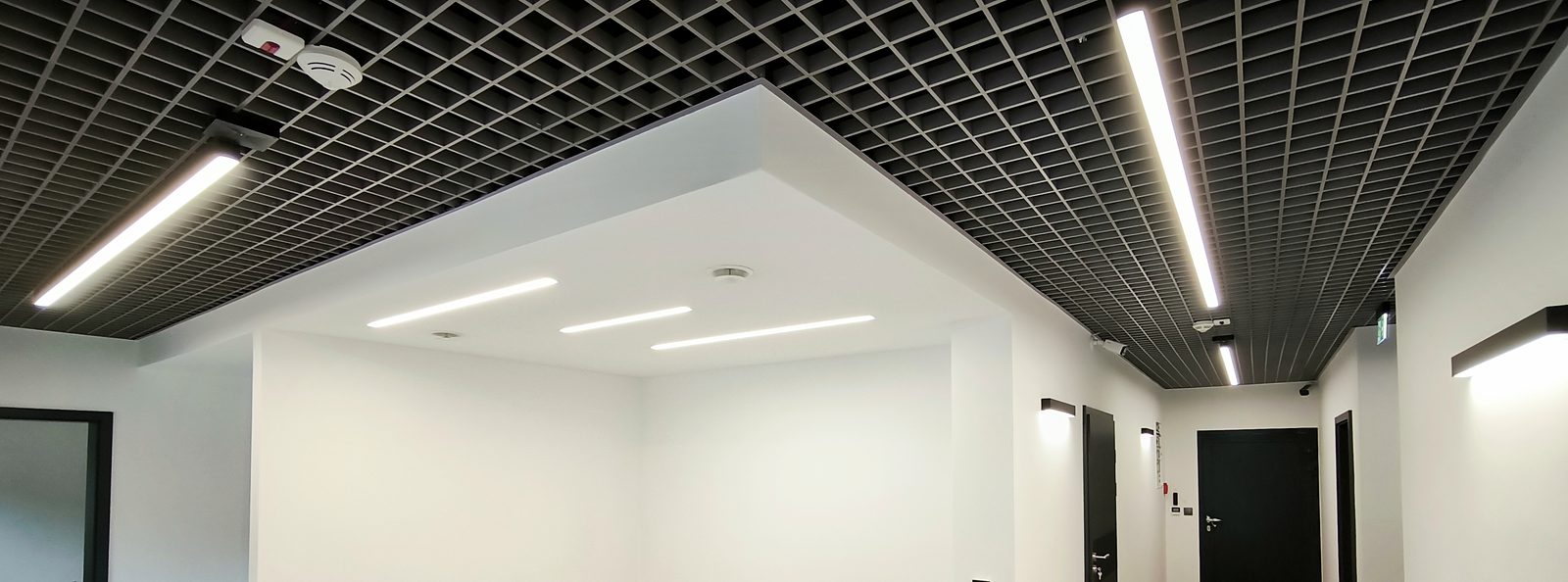 Hol budynku z liniami LED w suficie rastrowym i oprawami ściennymi